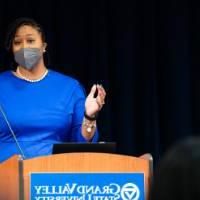 woman behind podium wearing mask speaks to audience, while gestering with her hands